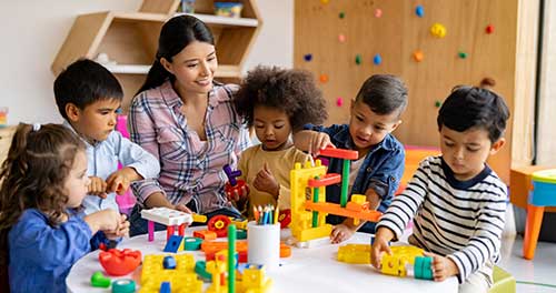 A front view of a childcare worker and some of her young pupils. They are playing with toy blocks in circle.