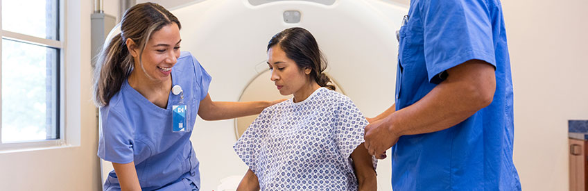 Two cheerful patient care technicians gently guide the mid adult woman onto the CT patient bed.